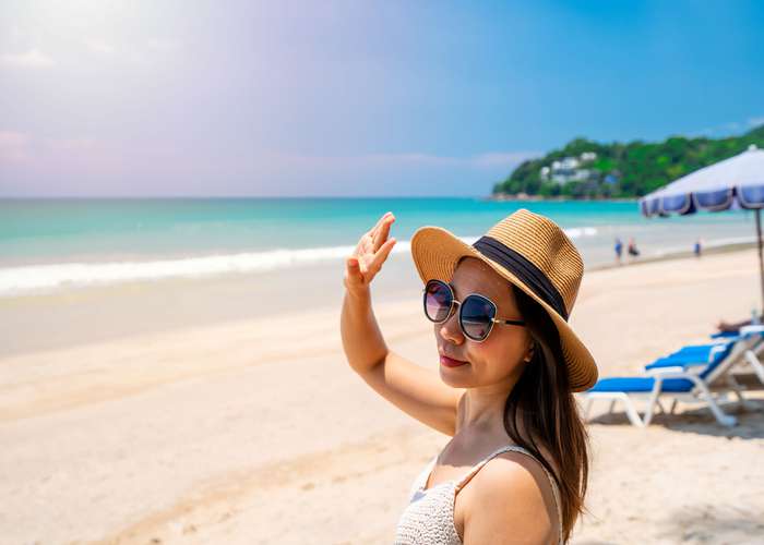 Woman standing in the sun. Wearing a hat and sunglasses, trying to cover her face from the sun with her hand.