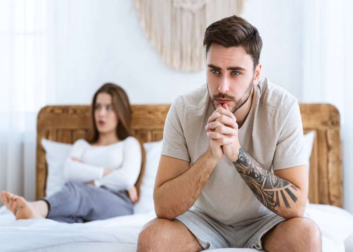 Man sitting on side of the bed feeling down because of erectile dysfunction, and woman laying in bed upset and unsatisfied.