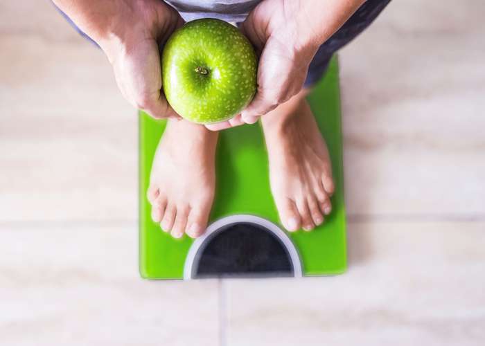 Maintain healthy weight for arthritis: person standing on a balance to measure his weight, holding a green apple.