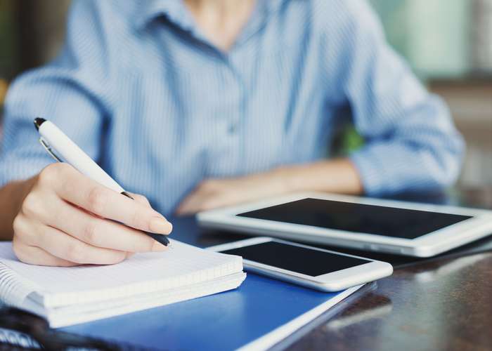 Female writing notes on a notebook with the pen to monitor her symptoms. Phone and Ipad next to her.