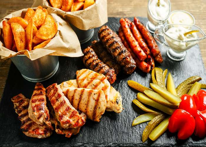Big plate of food consisting of grilled chicken, meat, and hot dogs. On the side, fried potatoes, pickles, and garniture.